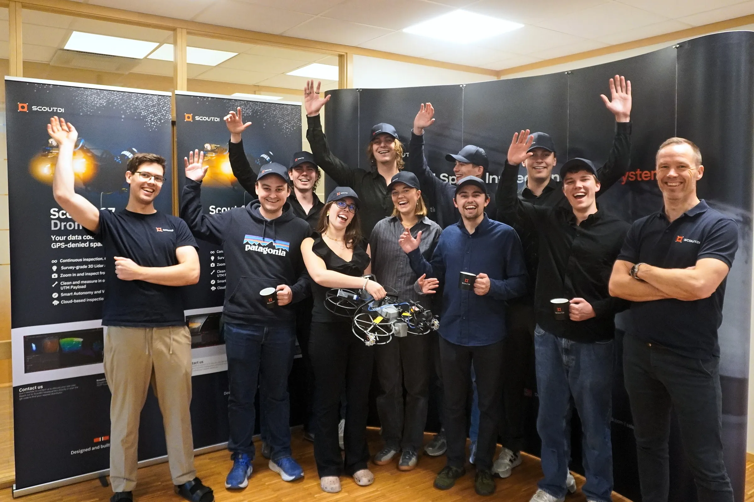 The Mobula team waving with ScoutDI in front of ScoutDI banners at their office in Trondheim, one of them holding a ScoutDI drone